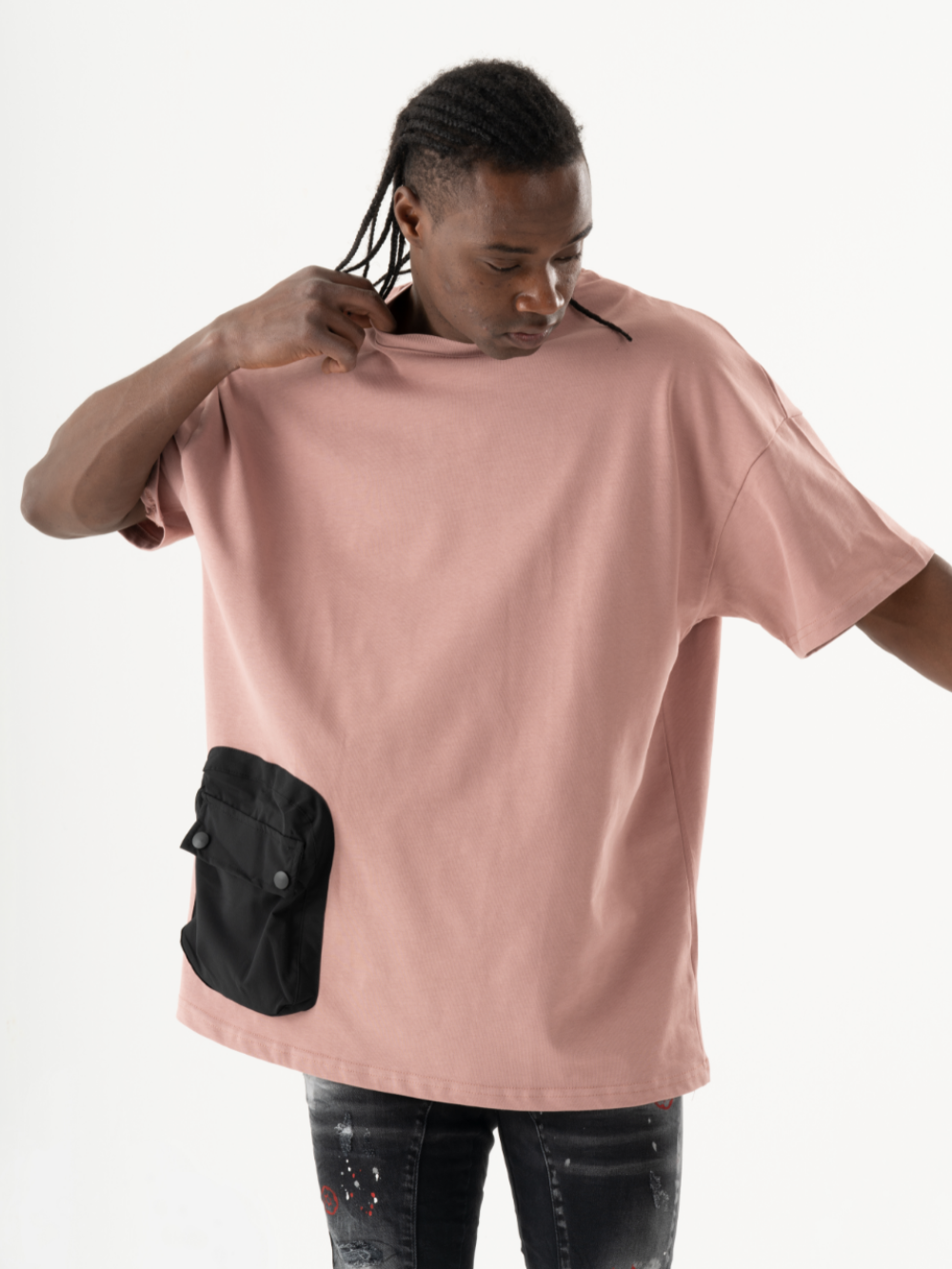 A person with braided hair wears a loose-fitting salmon short-sleeved DUNE T-SHIRT with a black buttoned front pocket and holds the neckline of the shirt with one hand. The background is white.