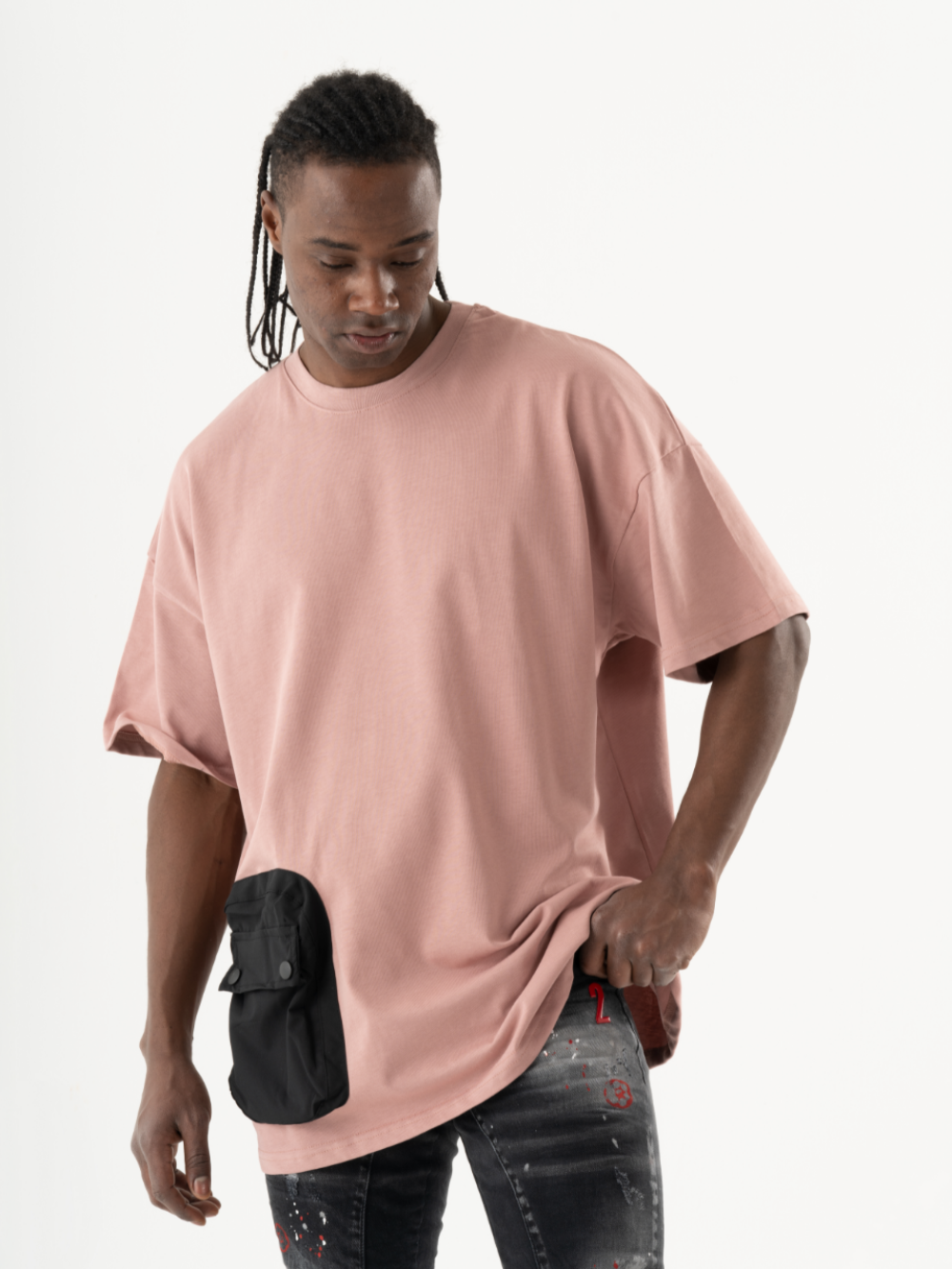 A person with long braids wearing a salmon DUNE T-SHIRT and black jeans with paint splatters stands looking down, with one hand holding a black pouch attached to the T-shirt.
