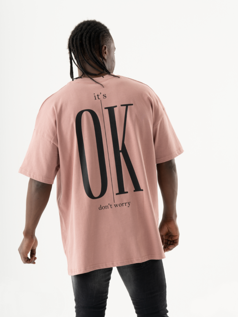 A person with long braided hair, wearing a pink oversized DUNE T-SHIRT with the words "it's OK don't worry" written on the back, stands with their back facing the camera.