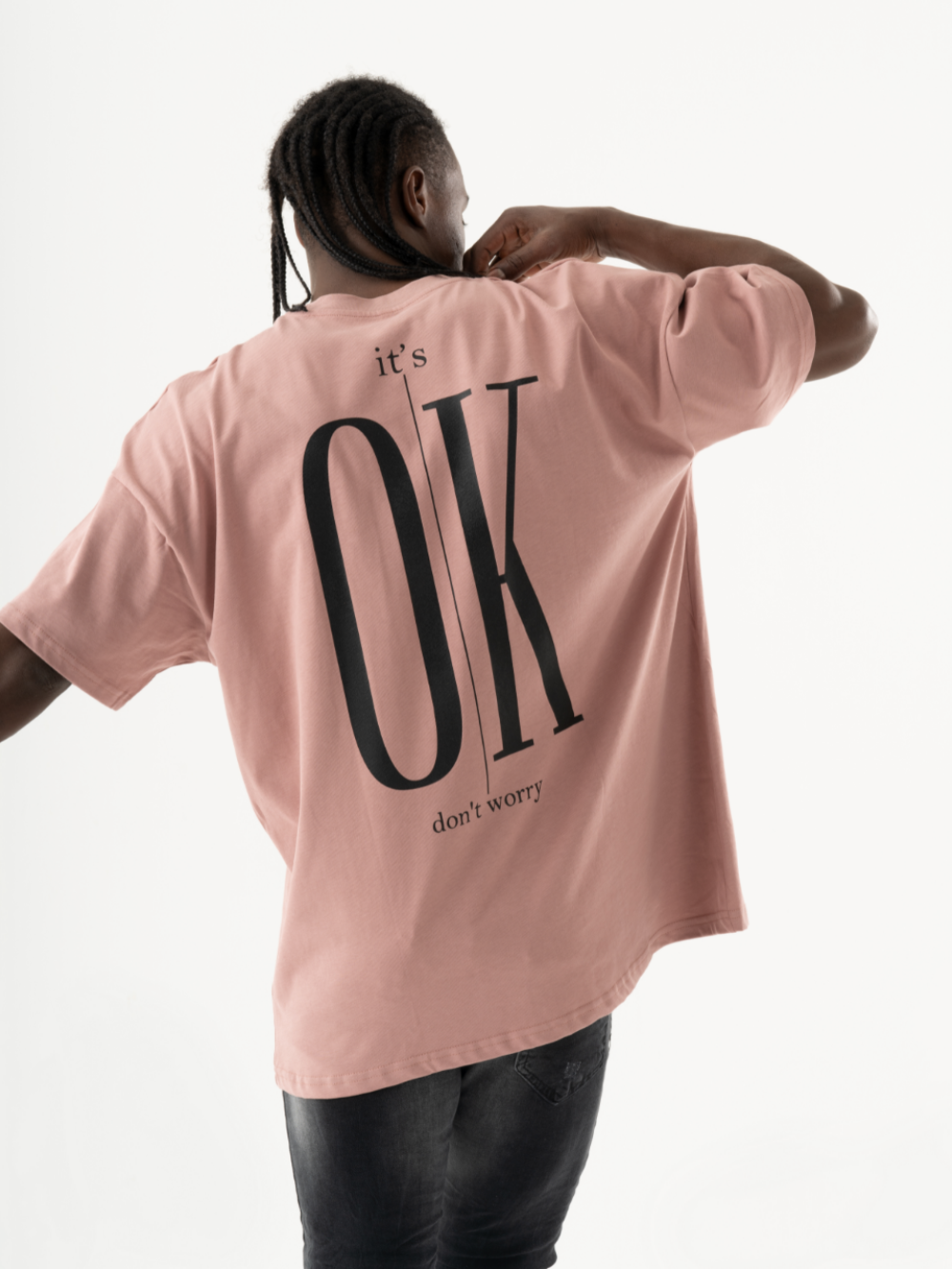 A person with braided hair is wearing a salmon DUNE T-SHIRT with the words "it's OK don't worry" printed on the back. The short-sleeved shirt, featuring a black buttoned front pocket, adds a touch of casual style as the individual faces away from the camera.