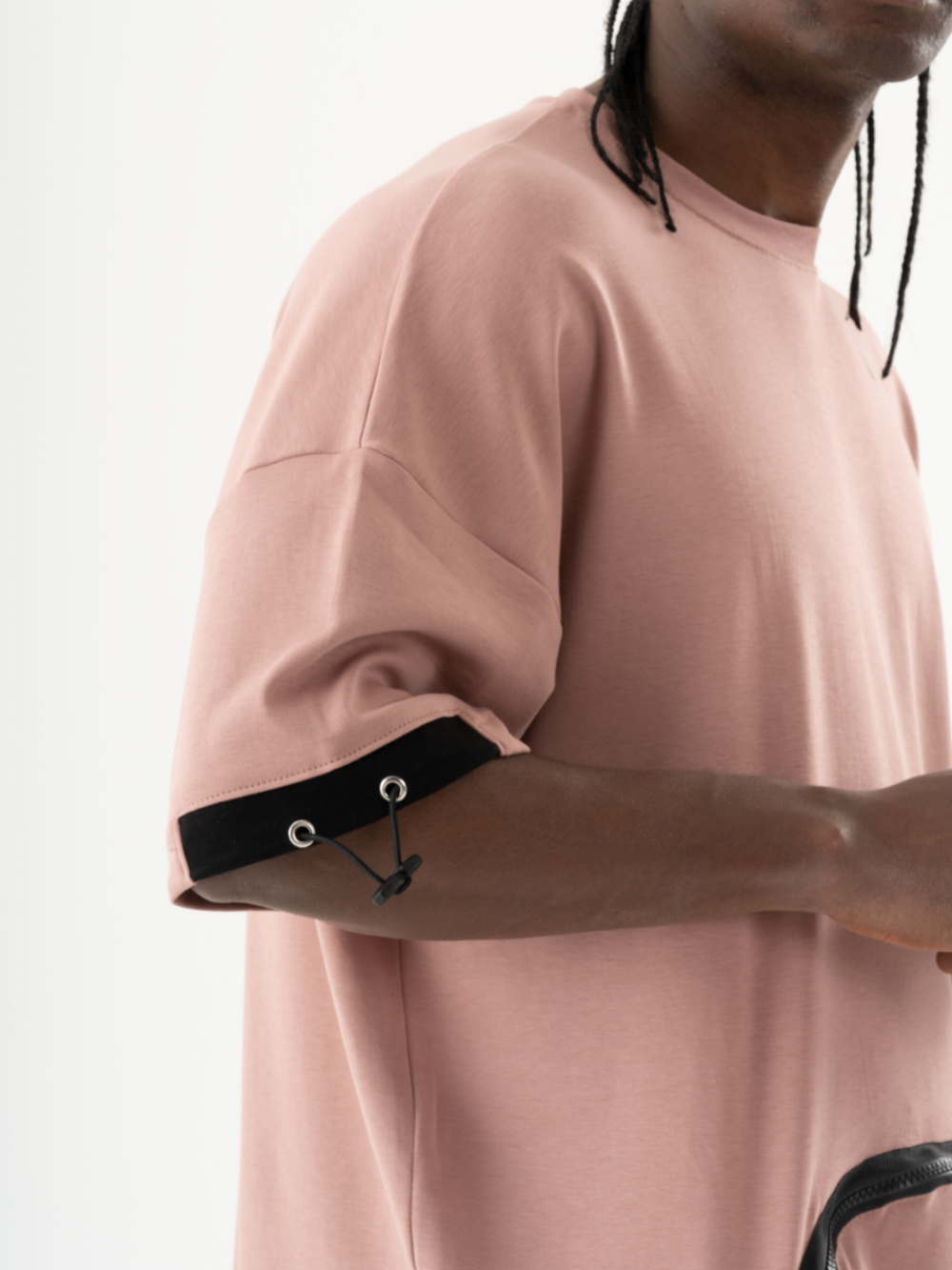 Man in a KANGAROO T-SHIRT with black drawstring details on the sleeves holding a black accessory. His head is turned, revealing braids. White background.