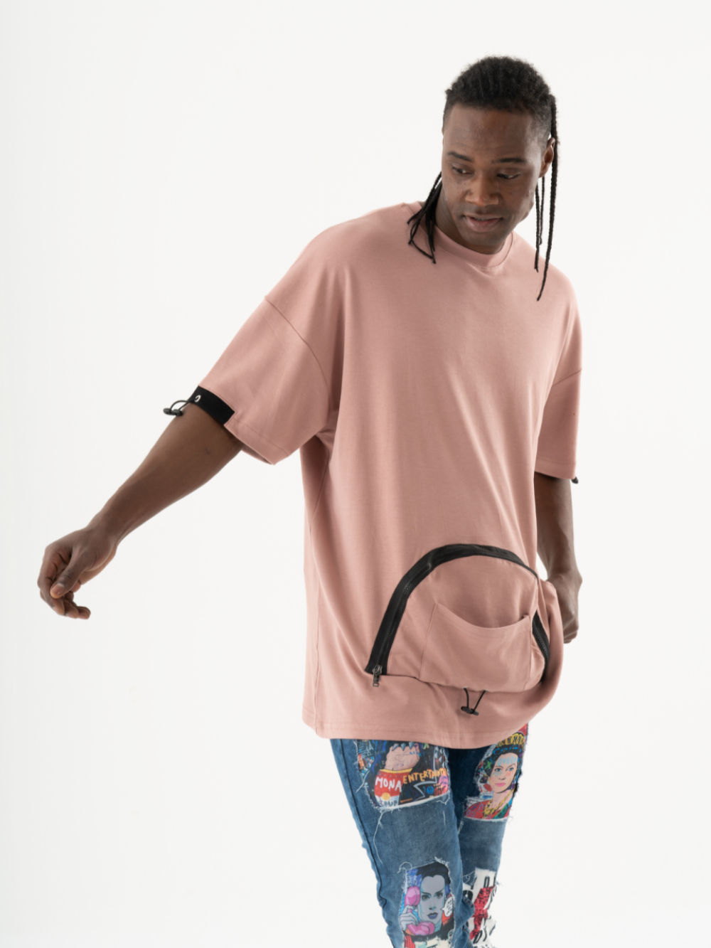 A man with braided hair wears a loose, salmon-colored KANGAROO T-SHIRT with a front pocket and decorative accents, paired with blue jeans featuring colorful patches. He looks down with his left arm slightly extended.