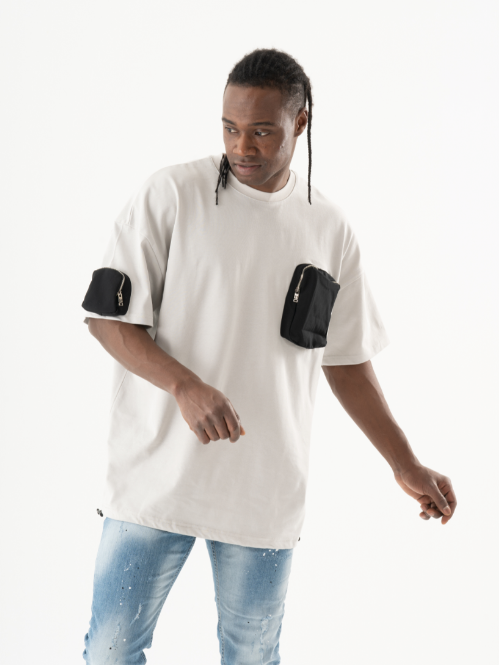 A man in a light beige oversized KLOUD T-SHIRT with black pockets on the sleeves, and blue jeans, effortlessly showcases a casual look against a plain white background.