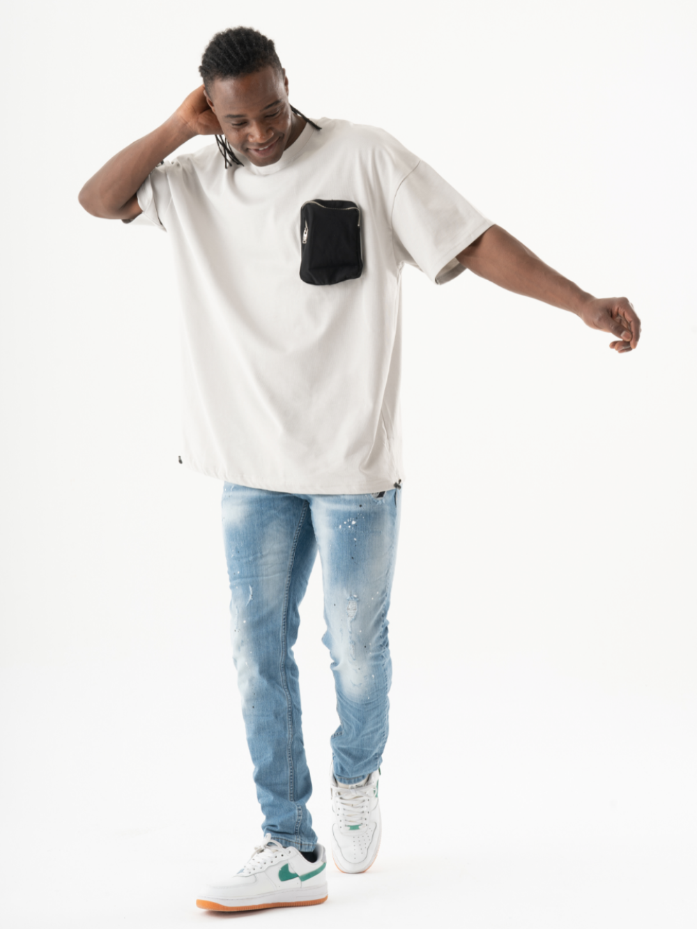 A person stands against a white background, epitomizing the casual look in a KLOUD T-SHIRT with black pockets, blue jeans, and white sneakers adorned with green accents. One hand touches their head while the other is extended outward.
