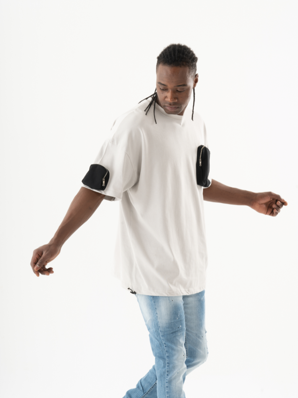A man with a casual look stands against a plain white background, wearing a KLOUD T-SHIRT and light blue jeans. His braided hair adds an element of style as he appears to be in motion, with his arms slightly extended.