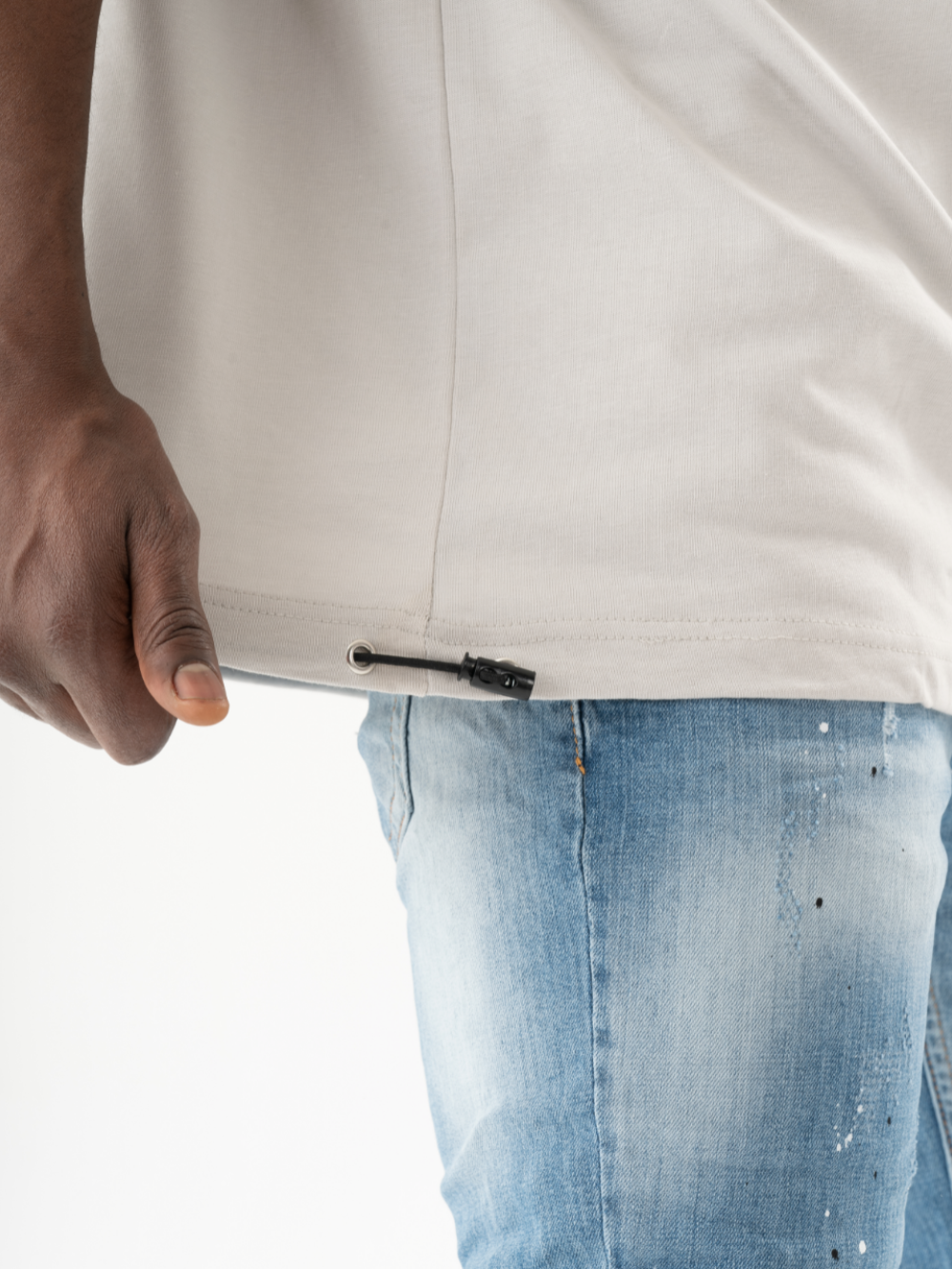 A person with a casual look is adjusting the drawstring at the bottom of a light-colored KLOUD T-SHIRT, while wearing blue jeans.