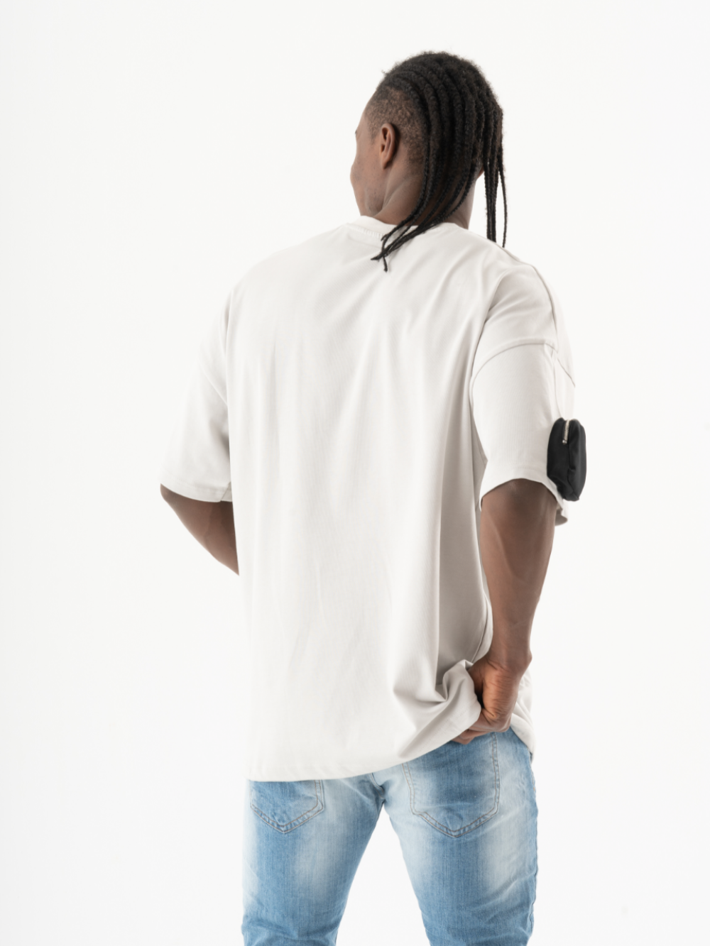 A man with braided hair, wearing a white KLOUD T-SHIRT, blue jeans, and a black armband, adopts a casual look as he stands facing away with his hands in his black pockets.