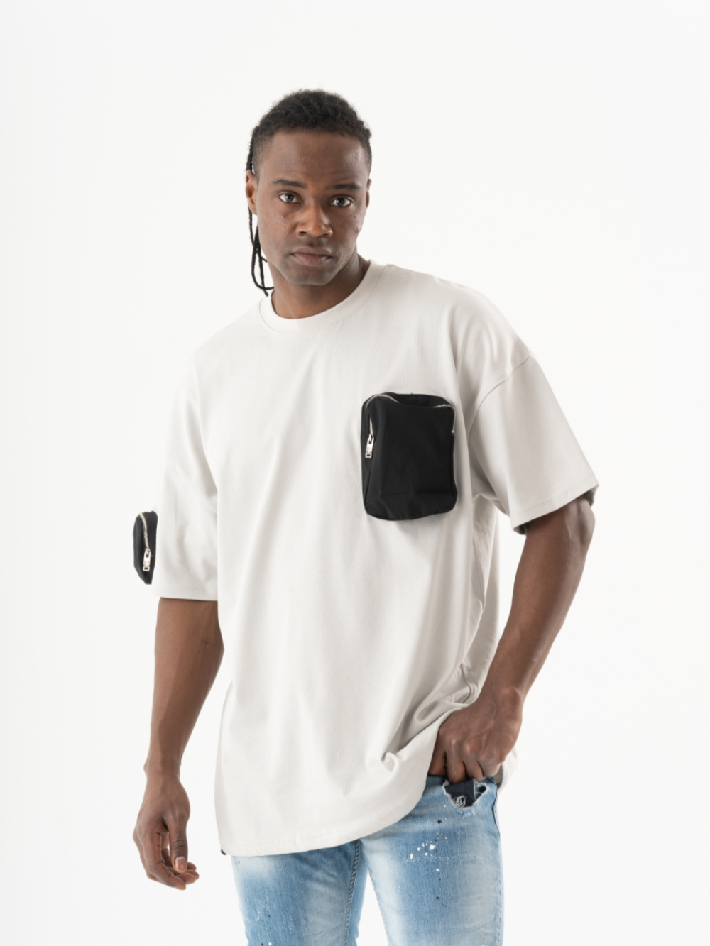 A man with a casual look, wearing a loose white KLOUD T-SHIRT with black zippered pockets on the chest and left sleeve, paired with light blue jeans. He has a serious expression and his left hand is in his pocket.