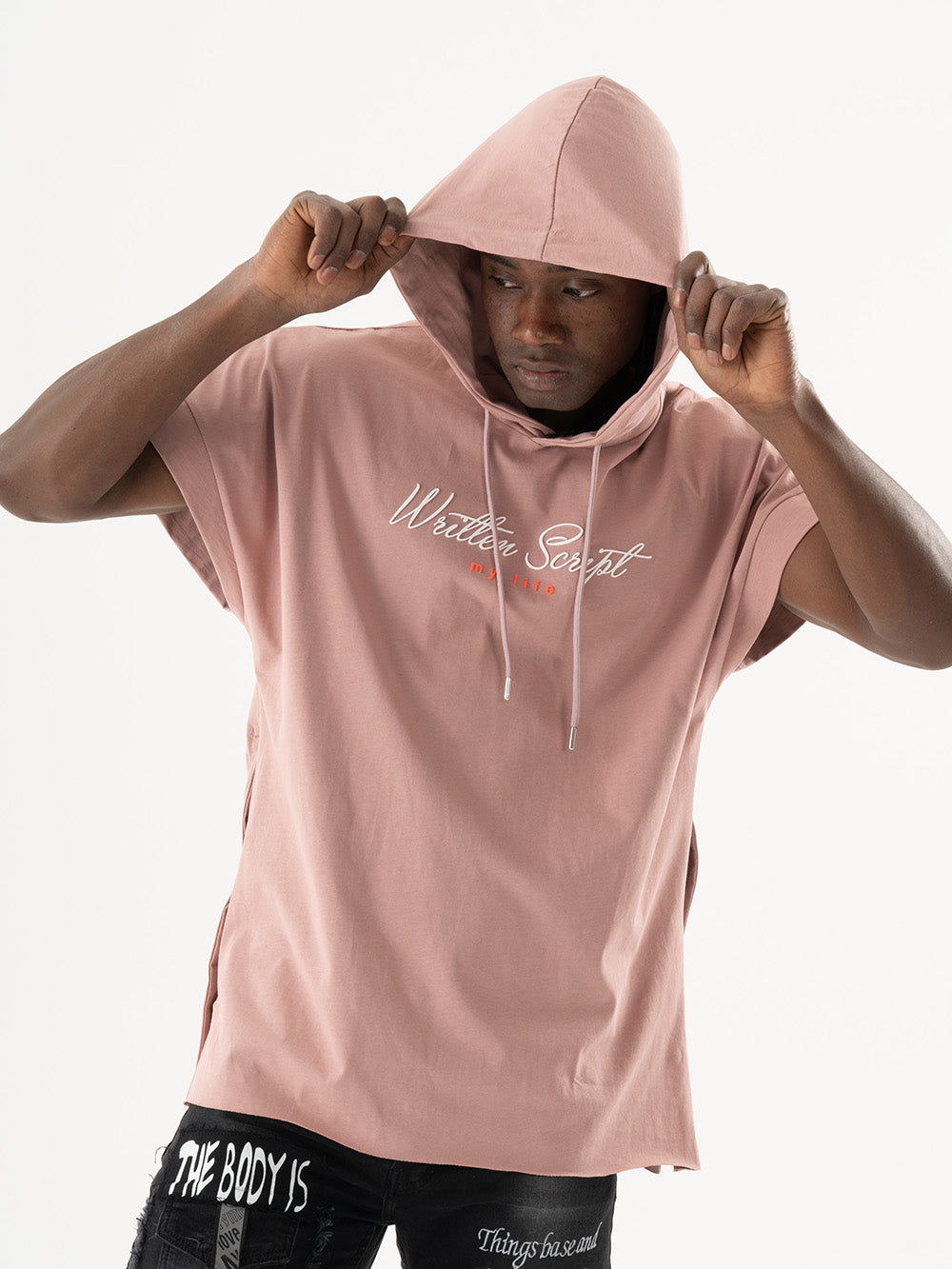 A man wearing a salmon short-sleeve RIGGED HOODIE with the text "Written Script" on the front, pulling the hood over his head. The comfort-fit hoodie matches his black pants adorned with white text patches, creating a casual and stylish look.