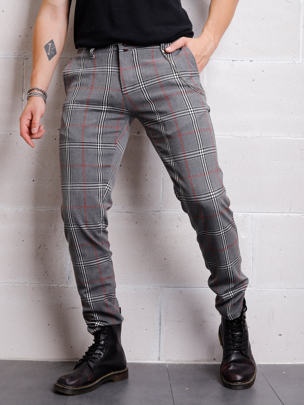 A man in a CHECKERED GREY shirt and black pants is leaning against a wall.