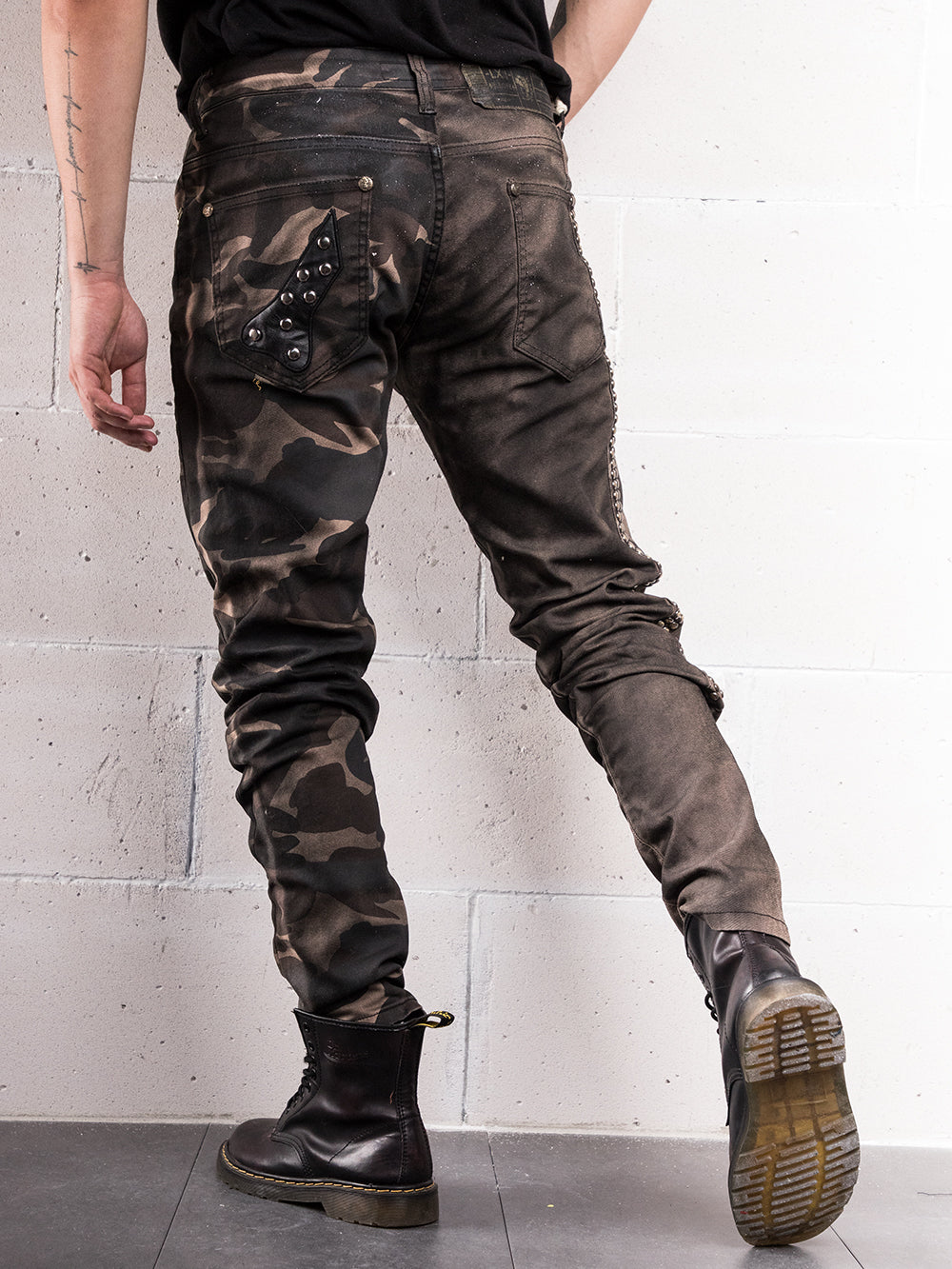A man wearing a pair of hand-crafted SNIPER camouflage jeans with elastic fabric, standing against a wall.
