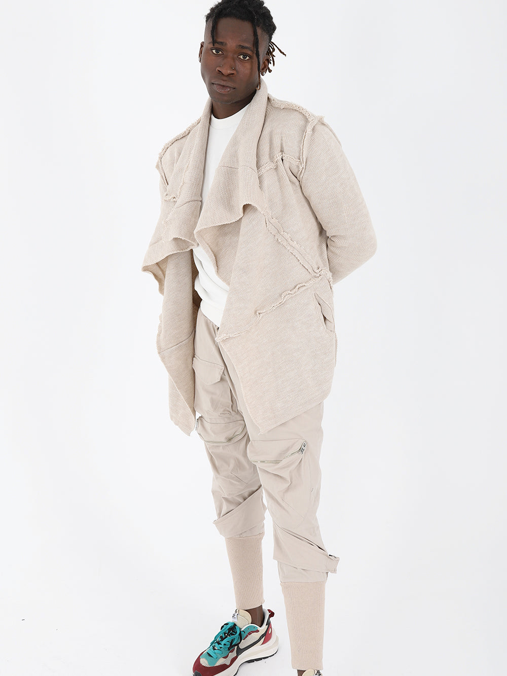 A man in a beige jacket and sneakers posing for a photo wearing the ASYMMETRIC SHORT CARDIGAN // IVORY, known for its true to size fit.