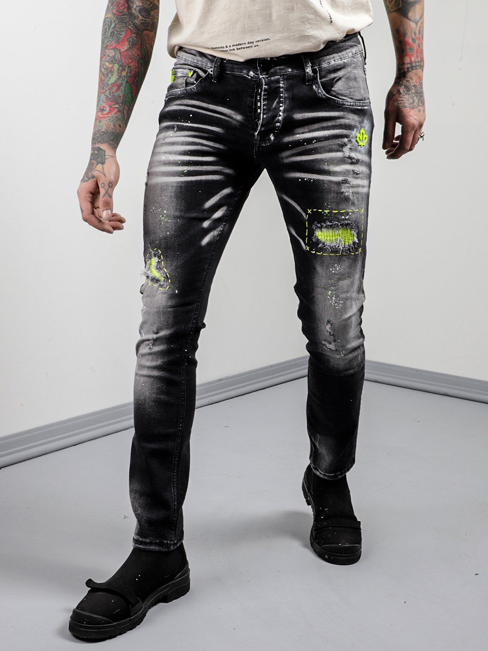 A man with tattoos and ripped jeans standing in front of a NEON TALK.