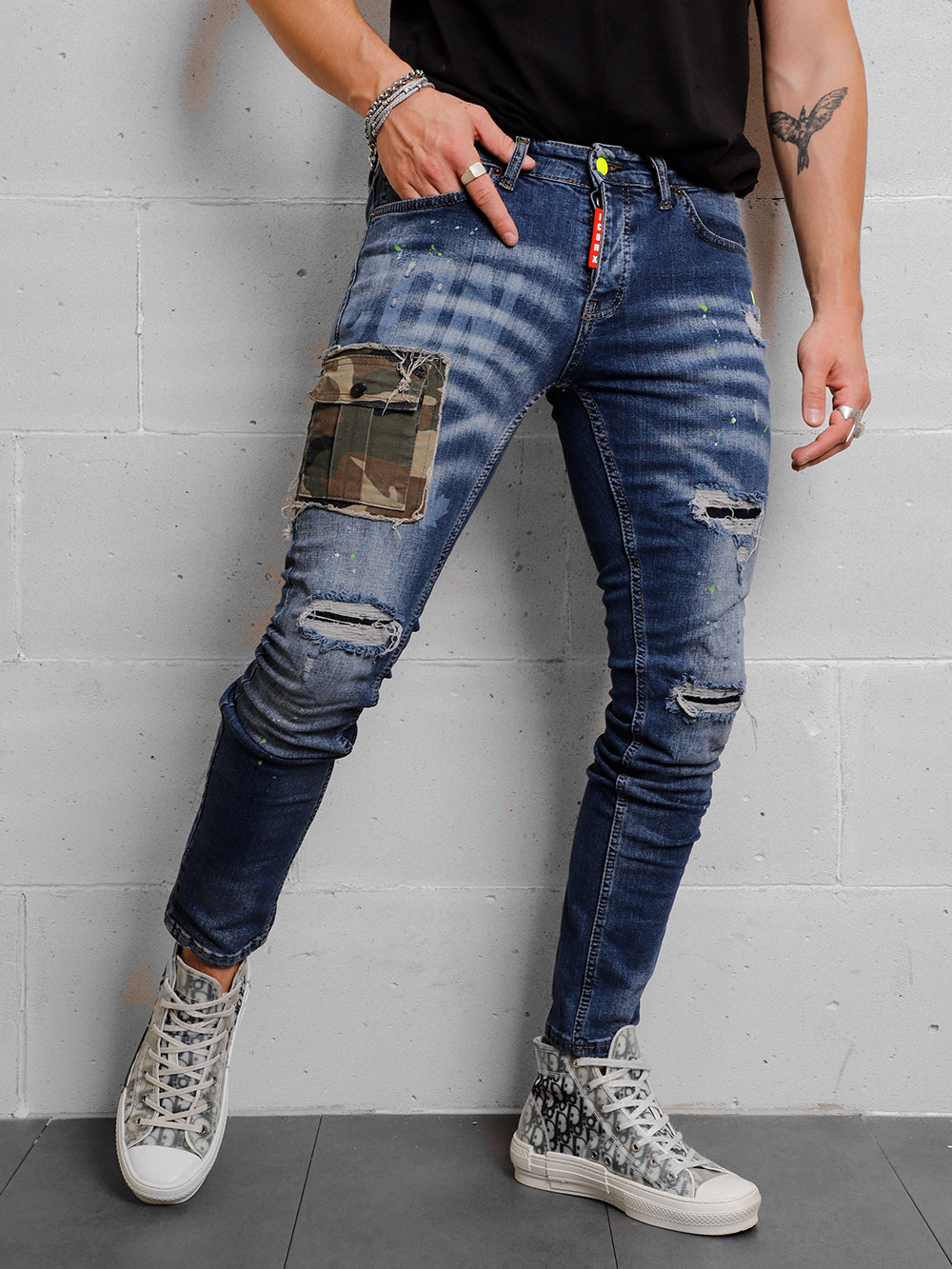 A man in a CAMOUFLAGE | BLUE t - shirt and jeans leaning against a wall.