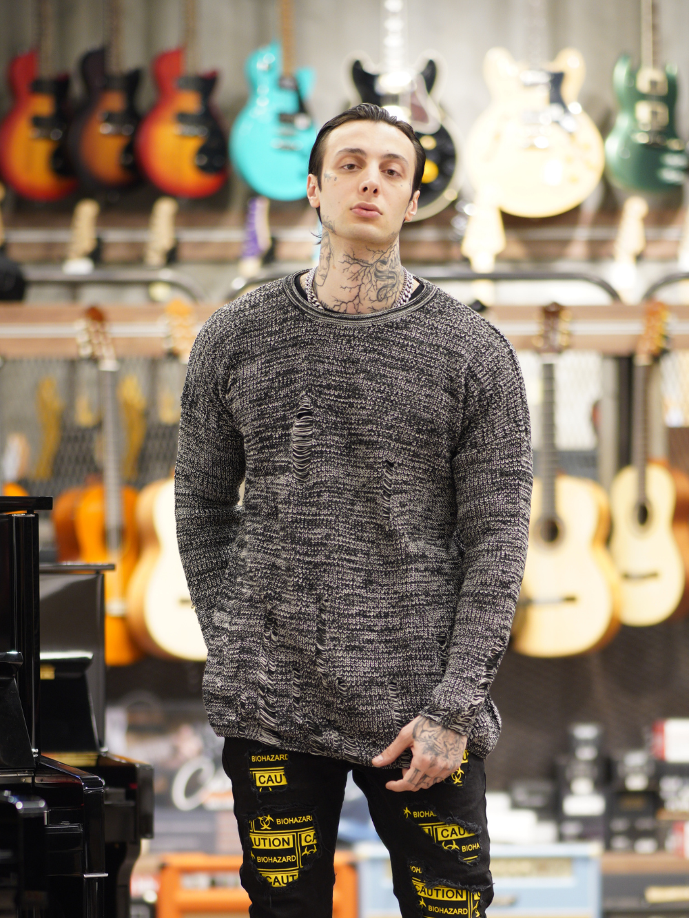 A man with a Distressed Gentleman Sweater | Gray standing in a room full of guitars.
