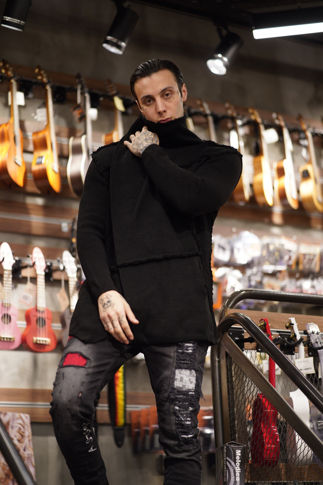 A man striking a pose in a store surrounded by guitars, showcasing the HOODED DISTRESSED CARDIGAN // BLACK and its diverse sizes and fits.