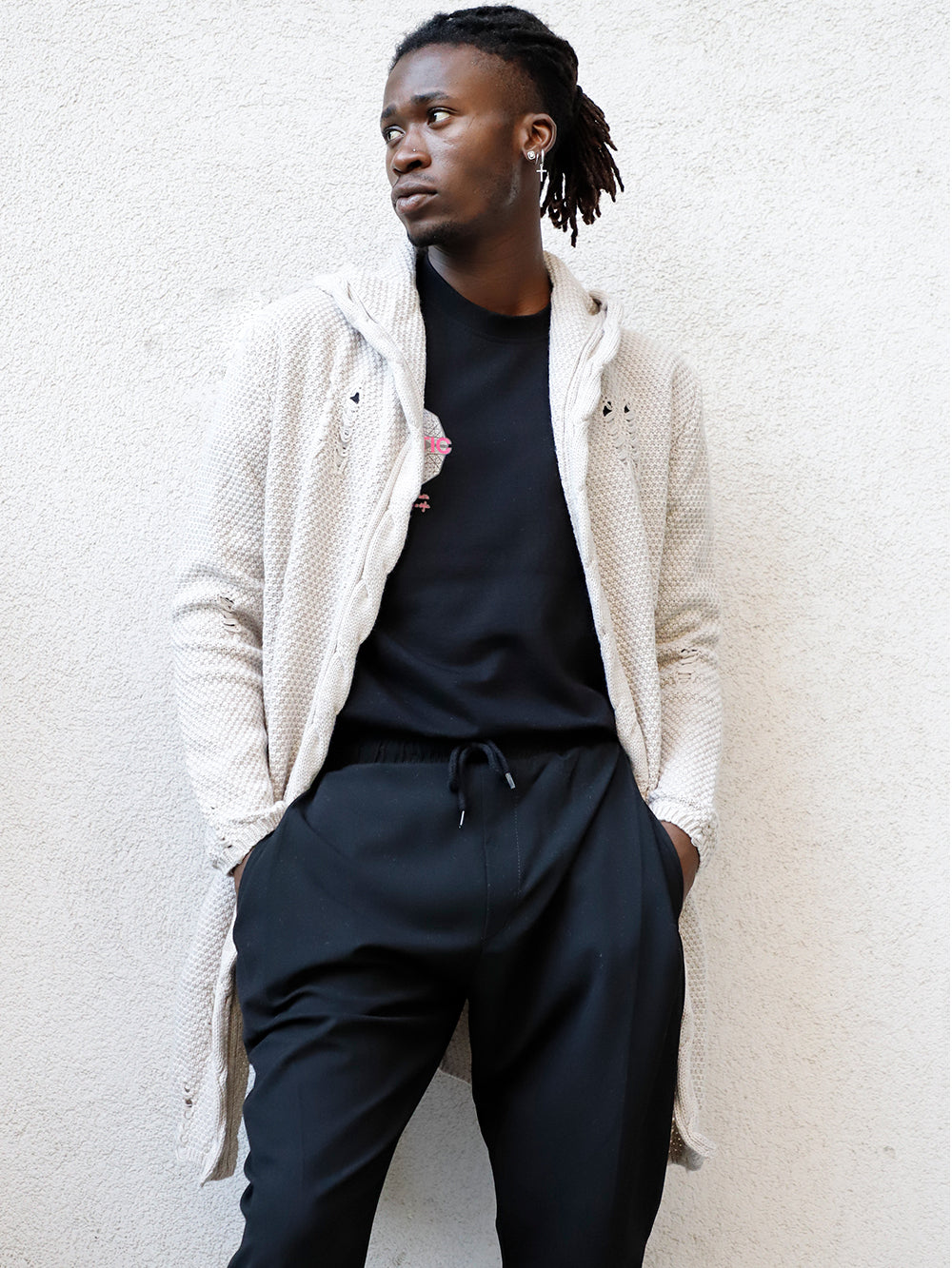 A black man wearing a HOODED DISTRESSED CARDIGAN // STONE leaning against a wall.