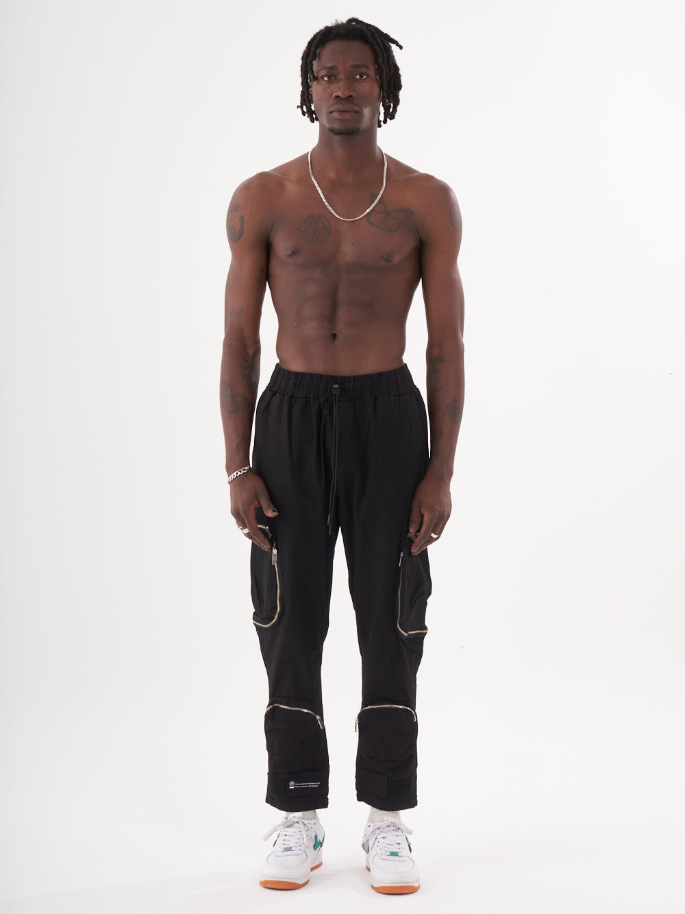 A shirtless man with dreadlocks stands facing forward, wearing the MULTI-ZIPPER POCKET JOGGER // BLACK for jogger comfort, paired with white sneakers.