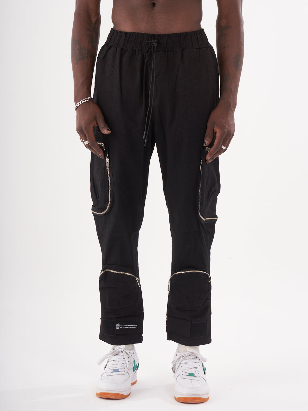 A person wearing MULTI-ZIPPER POCKET JOGGER // BLACK with an elastic drawstring waist for jogger comfort, white sneakers, and a black bracelet stands against a plain white background.