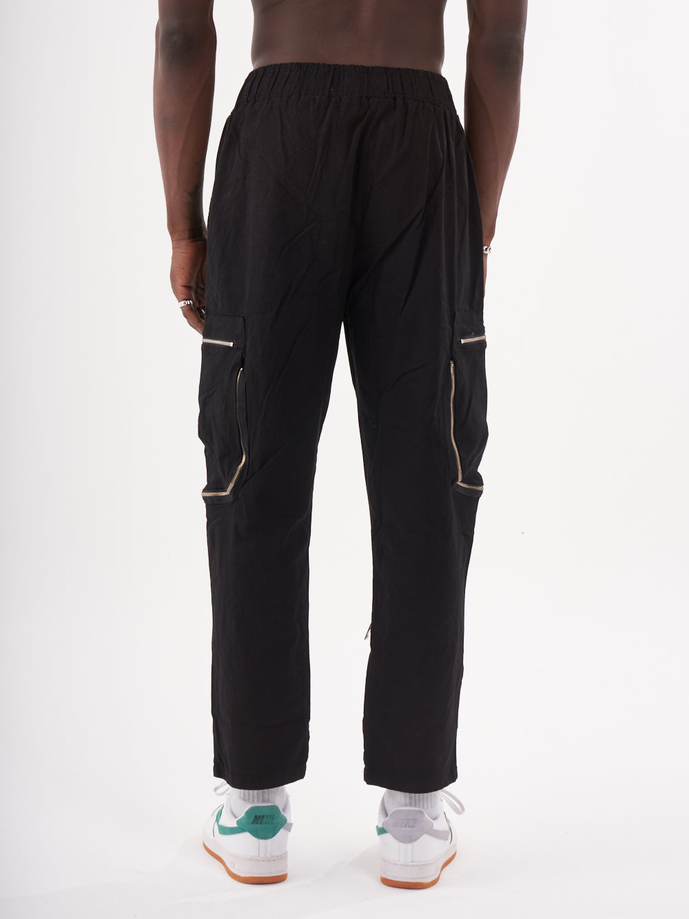 A person is wearing MULTI-ZIPPER POCKET JOGGER // BLACK, featuring a slim fit and an elastic drawstring waist for jogger comfort. They stand against a plain white background, also dressed in white socks and white sneakers with green accents.