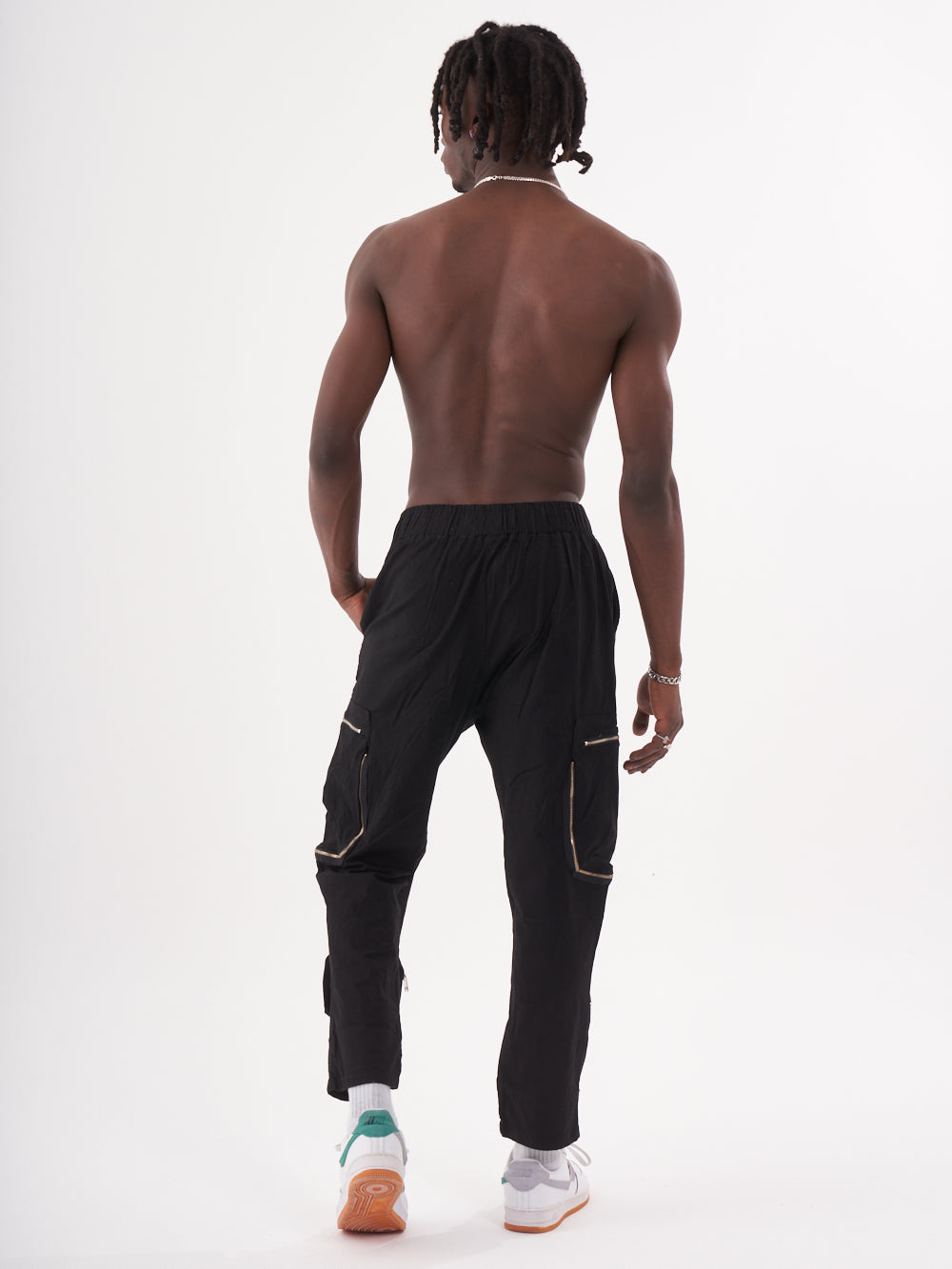 A shirtless person with braided hair stands facing away, wearing MULTI-ZIPPER POCKET JOGGER // BLACK and white sneakers with a green design on the soles.