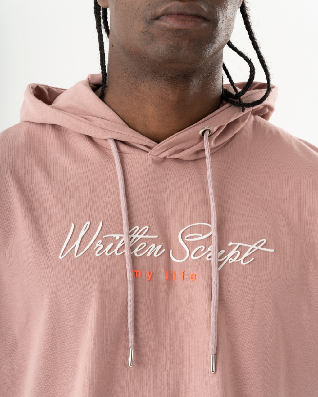 Close-up of a person wearing a light pink, comfort fit RIGGED HOODIE with "Written Script" and "my life" embroidered on the front in white and red text. The salmon short sleeve hoodie has button closure side slits, adding a unique touch. The person has braided hair.