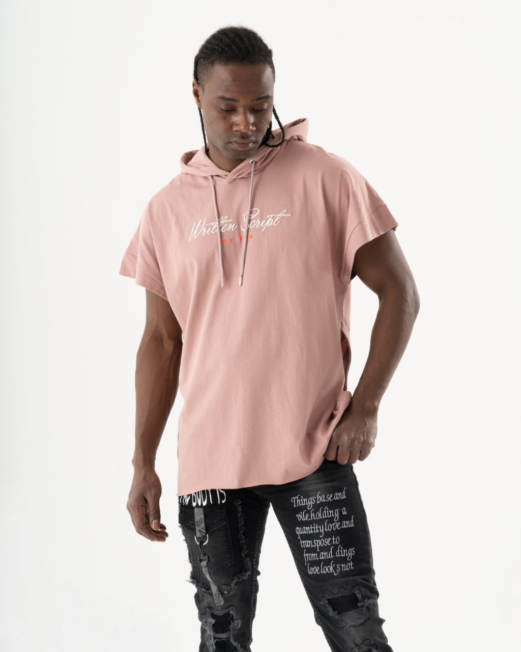 A person wearing a salmon RIGGED HOODIE and black jeans with white text printed on them, standing against a white background and looking down. The comfort fit of the attire exudes a relaxed vibe.