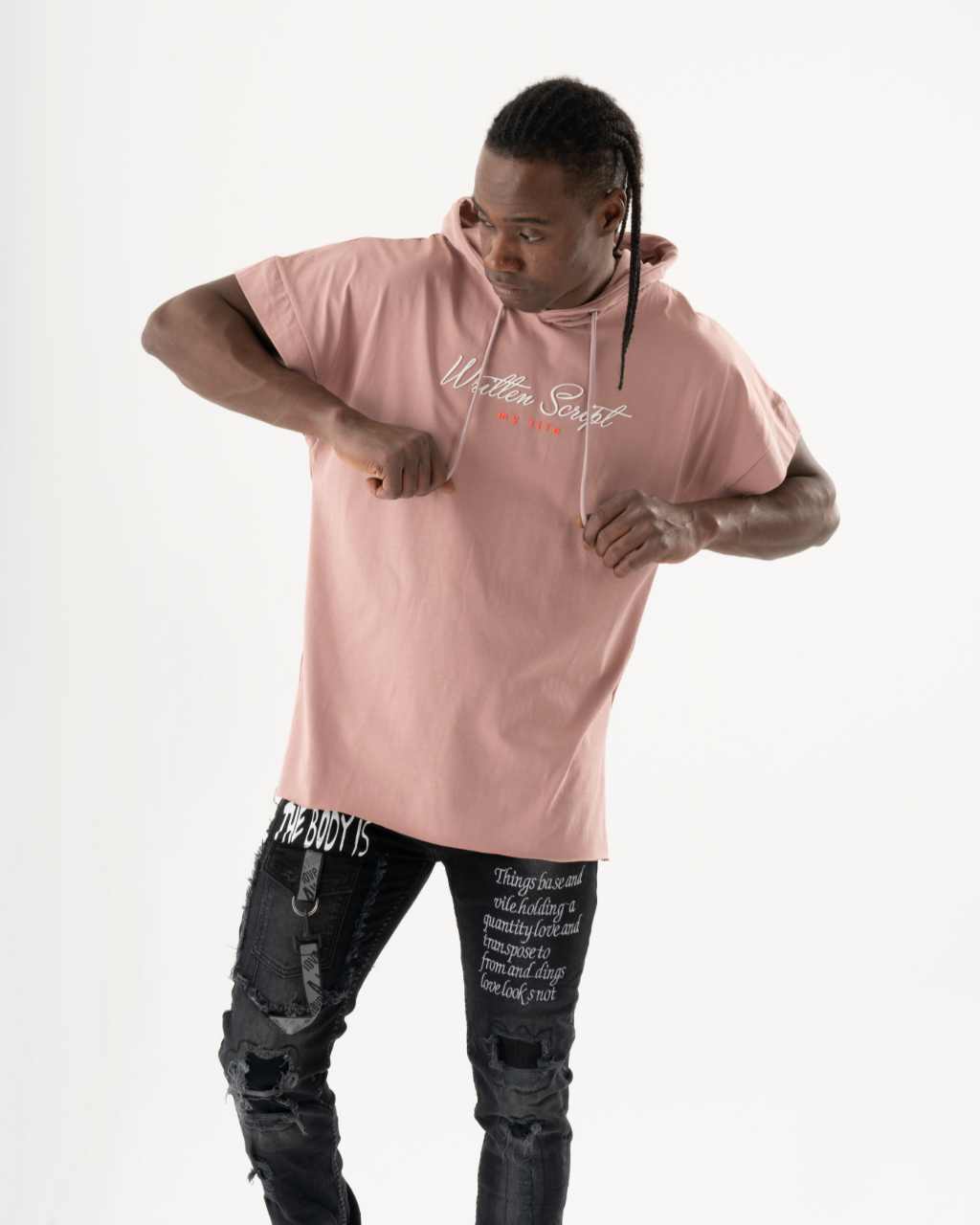 A man wearing a salmon RIGGED HOODIE and black pants with white text strikes a pose. He has long braided hair and looks down with a serious expression. The background is plain white, highlighting the comfort fit of his attire.