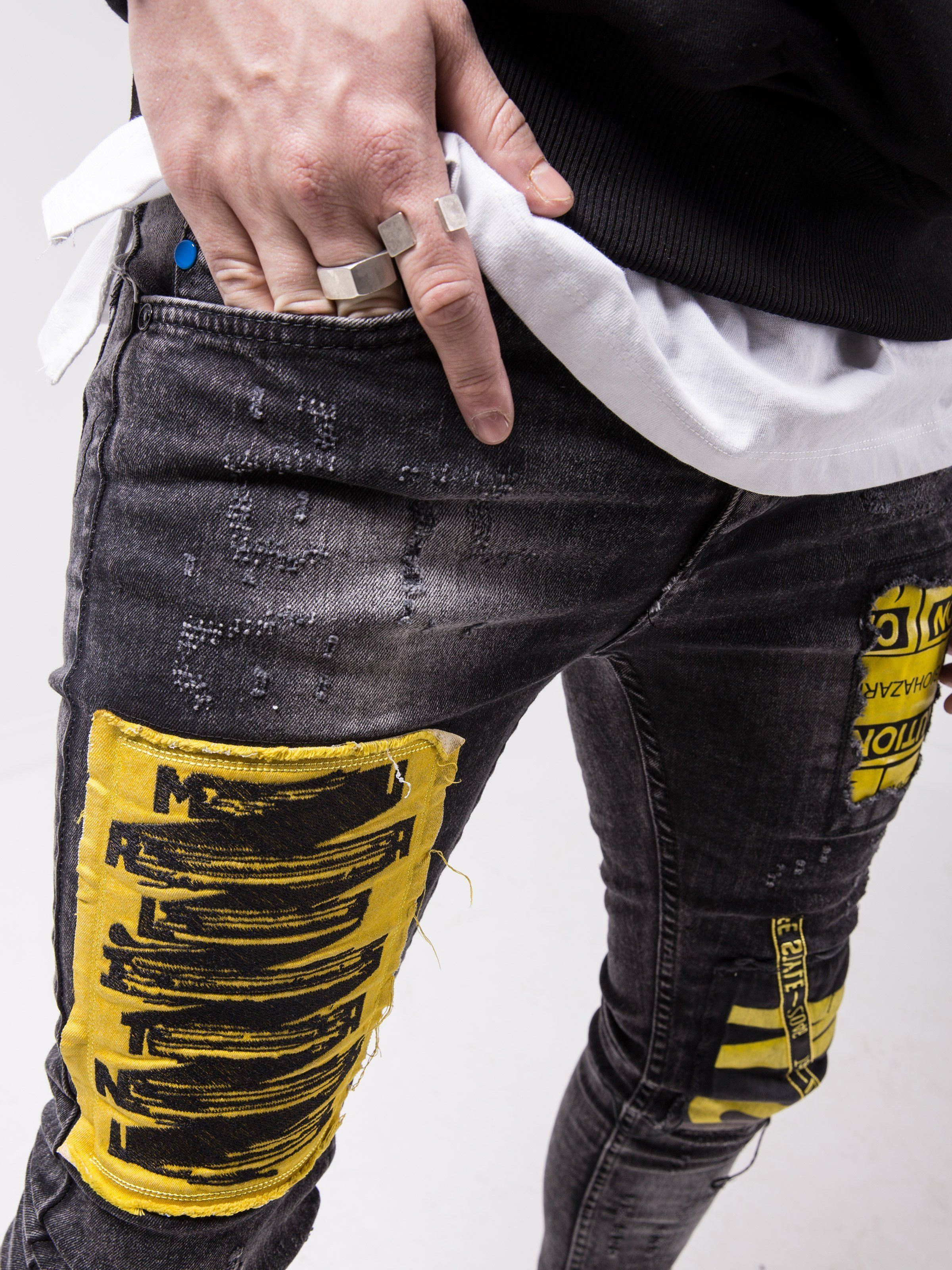A man wearing MOUNT ONE skinny fit black jeans with yellow patches on them.