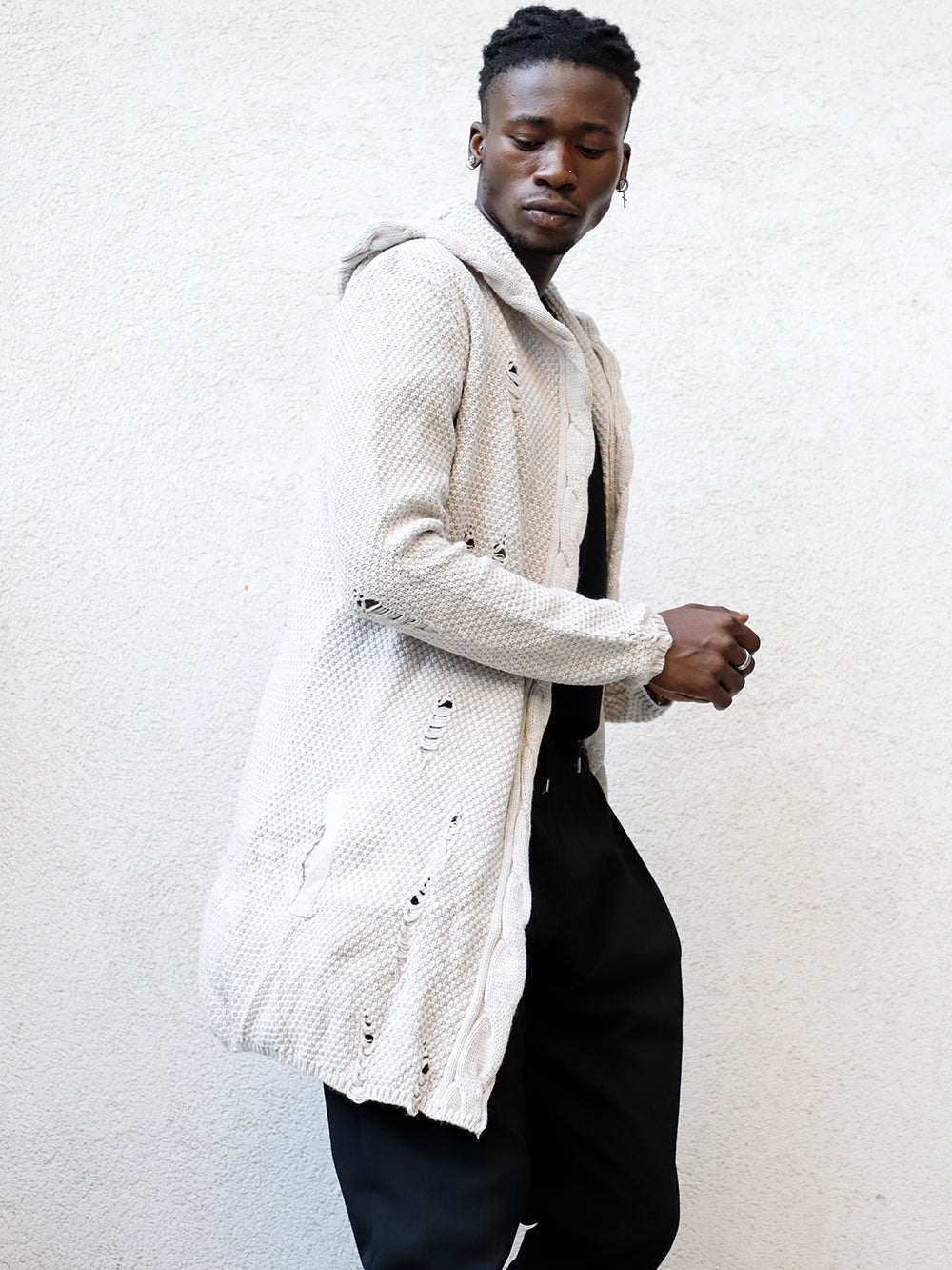 A man in Hooded Distressed Cardigan // Stone leaning against a wall.