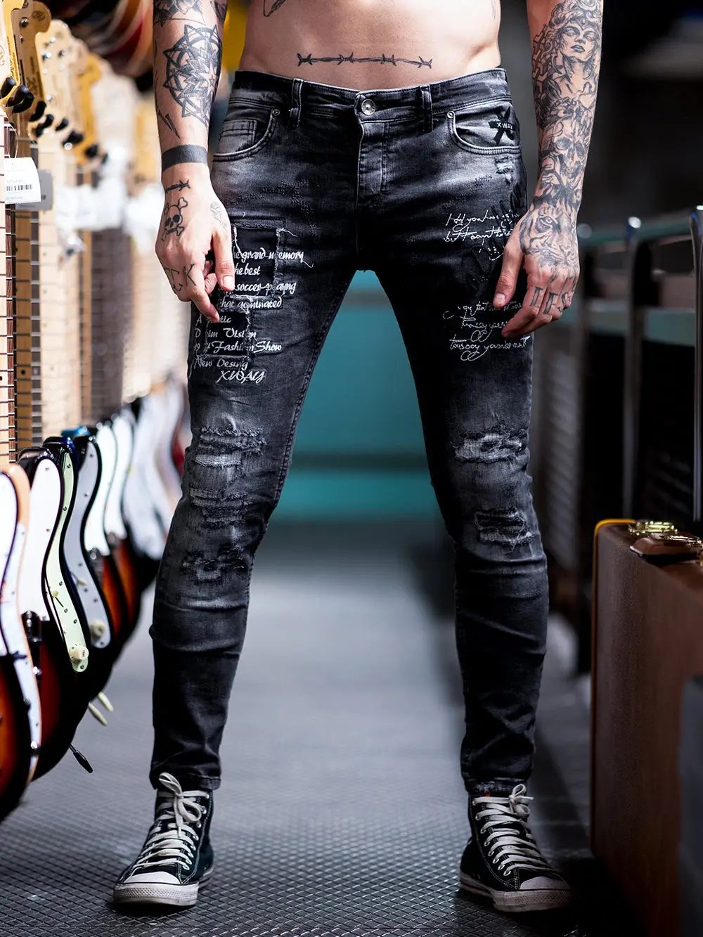 Photo of the lower half of a man with tattoos standing wearing BLACK STONE jeans by SERNES. The distressed jeans has embroidered text on it.