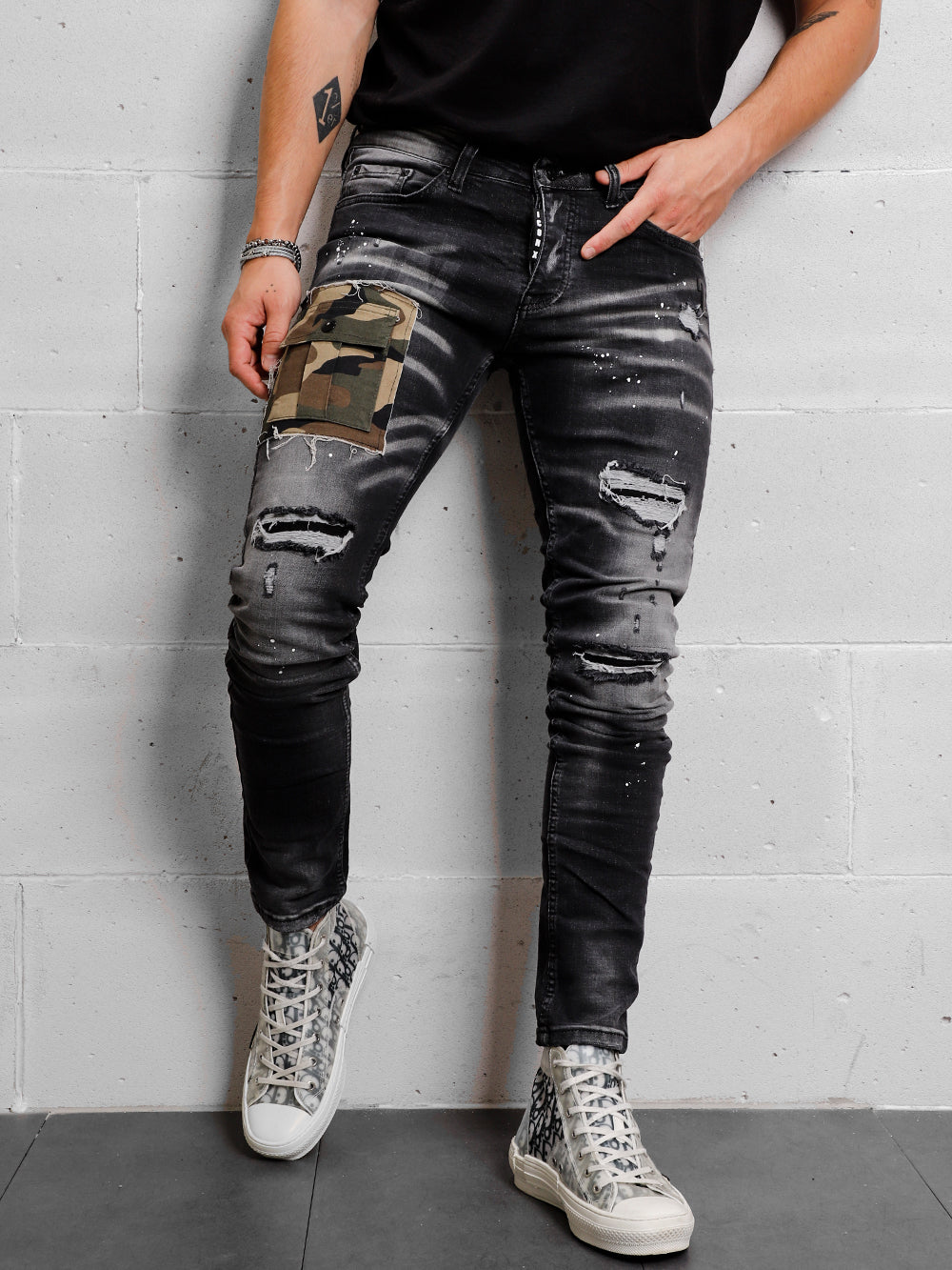 A man in a CAMOUFLAGE | BLACK t - shirt and jeans leaning against a wall.