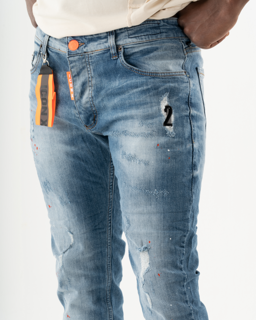 A man is wearing a pair of TEZAR with an orange tag. The jeans have patches and a skinny fit.