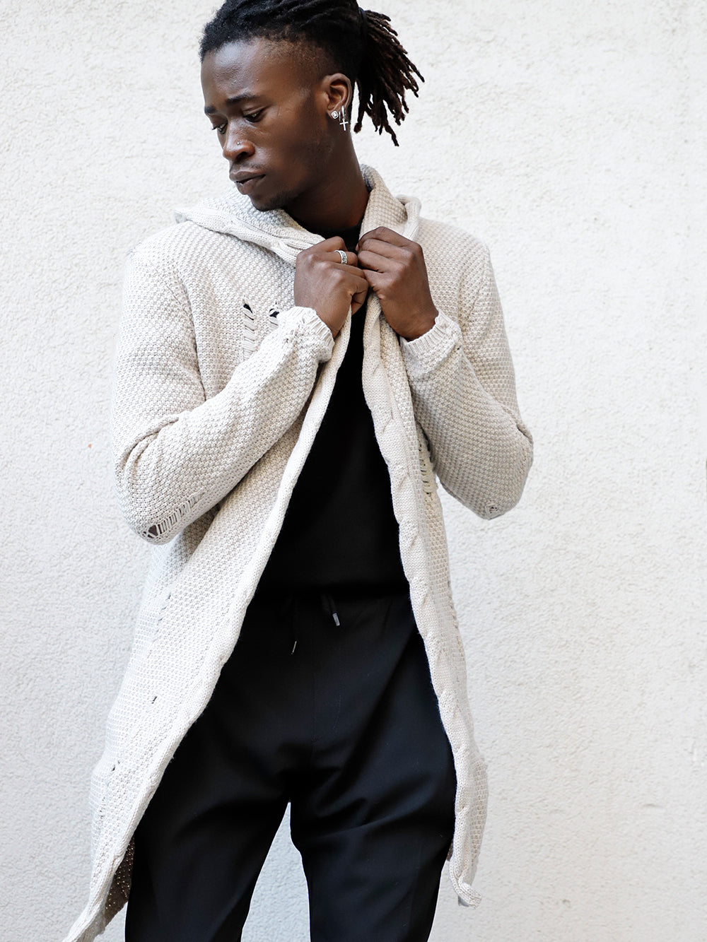 A man in hooded distressed cardigan // stone skinny fit jeans and a white jacket leaning against a wall.