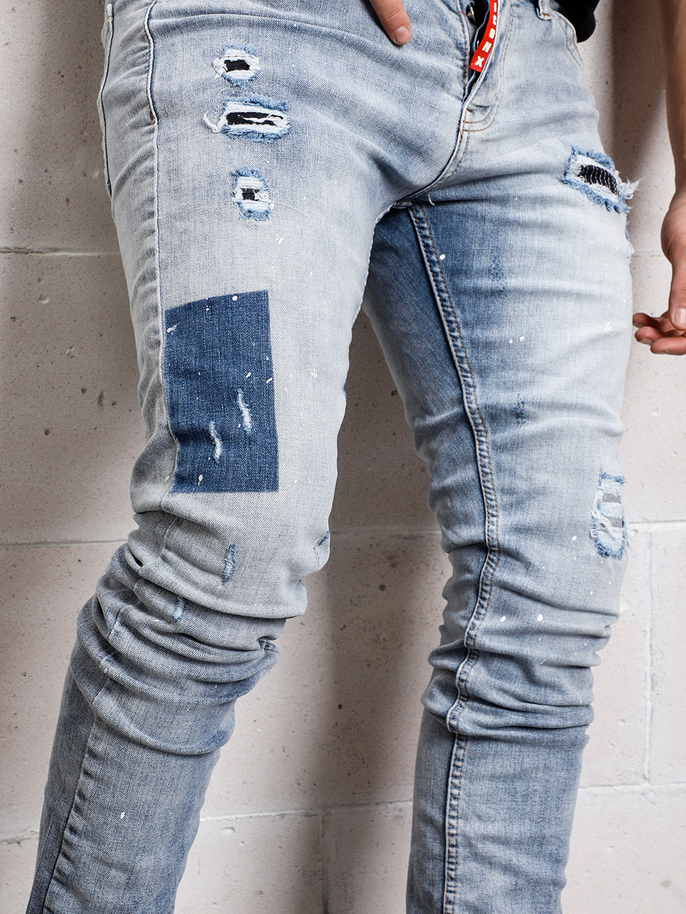 A man wearing ripped jeans with distressed details, leaning against a wall in his SKY BLUE ICON DENIM.