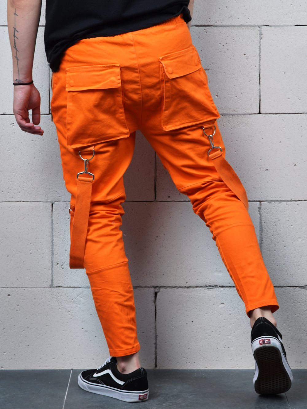 A man wearing ORANGE BRONX cargo pants is leaning against a brick wall.