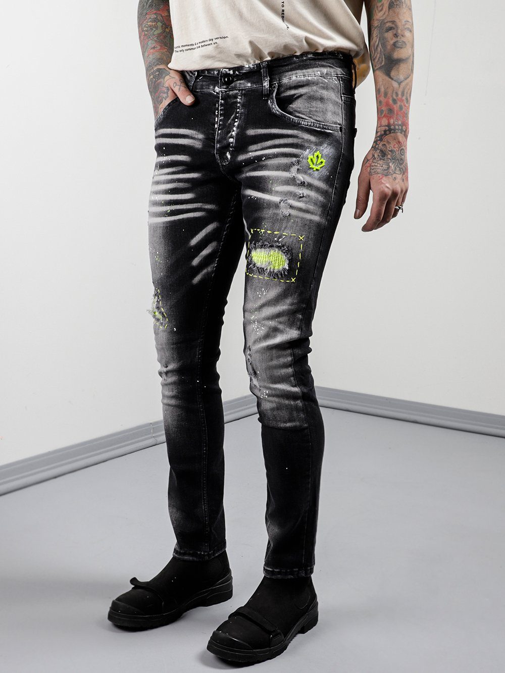 A man wearing NEON TALK and ripped jeans is standing in front of a white wall.