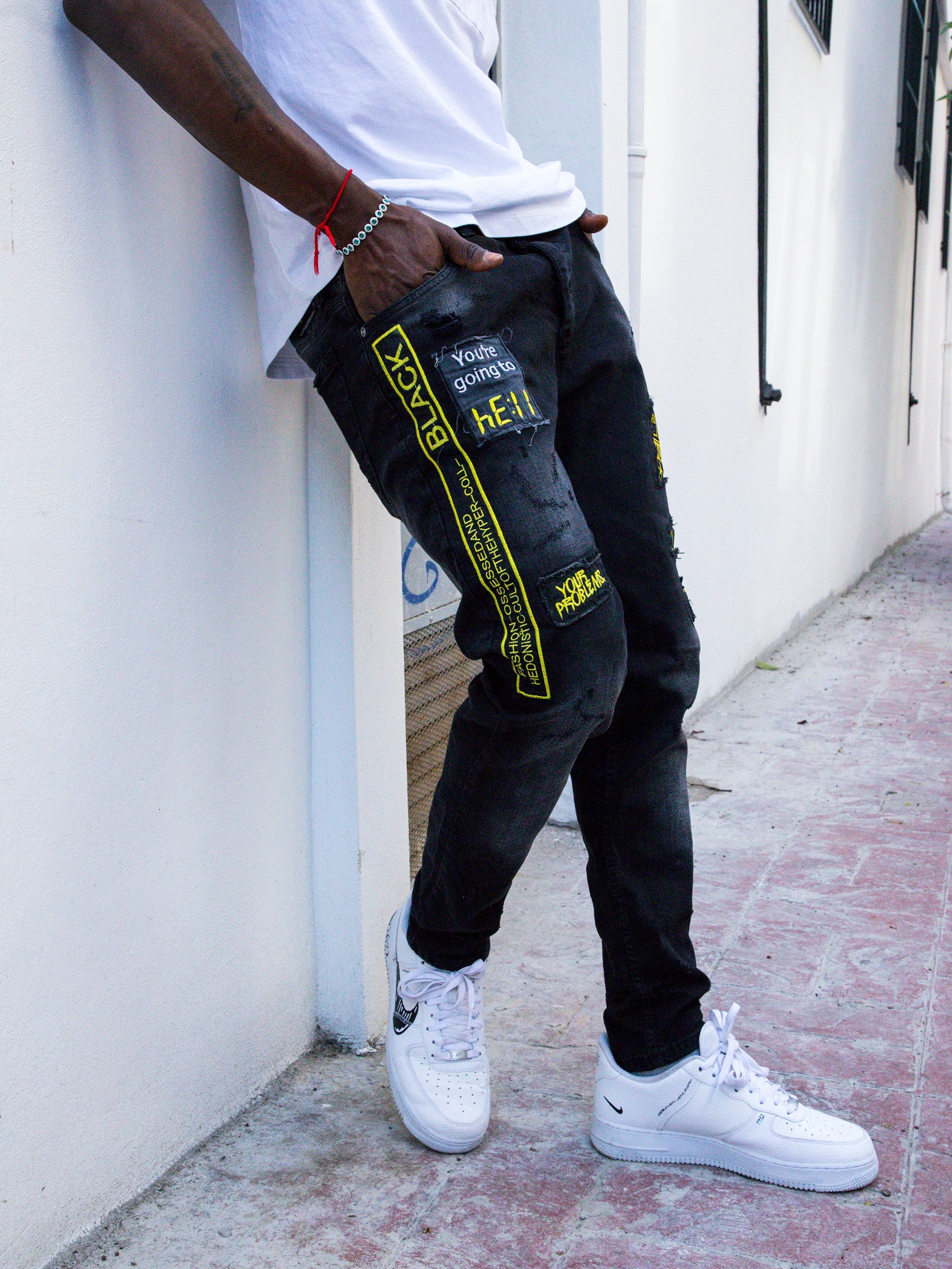 A man leaning against a wall wearing BLACK FALCON jeans and a white t-shirt.