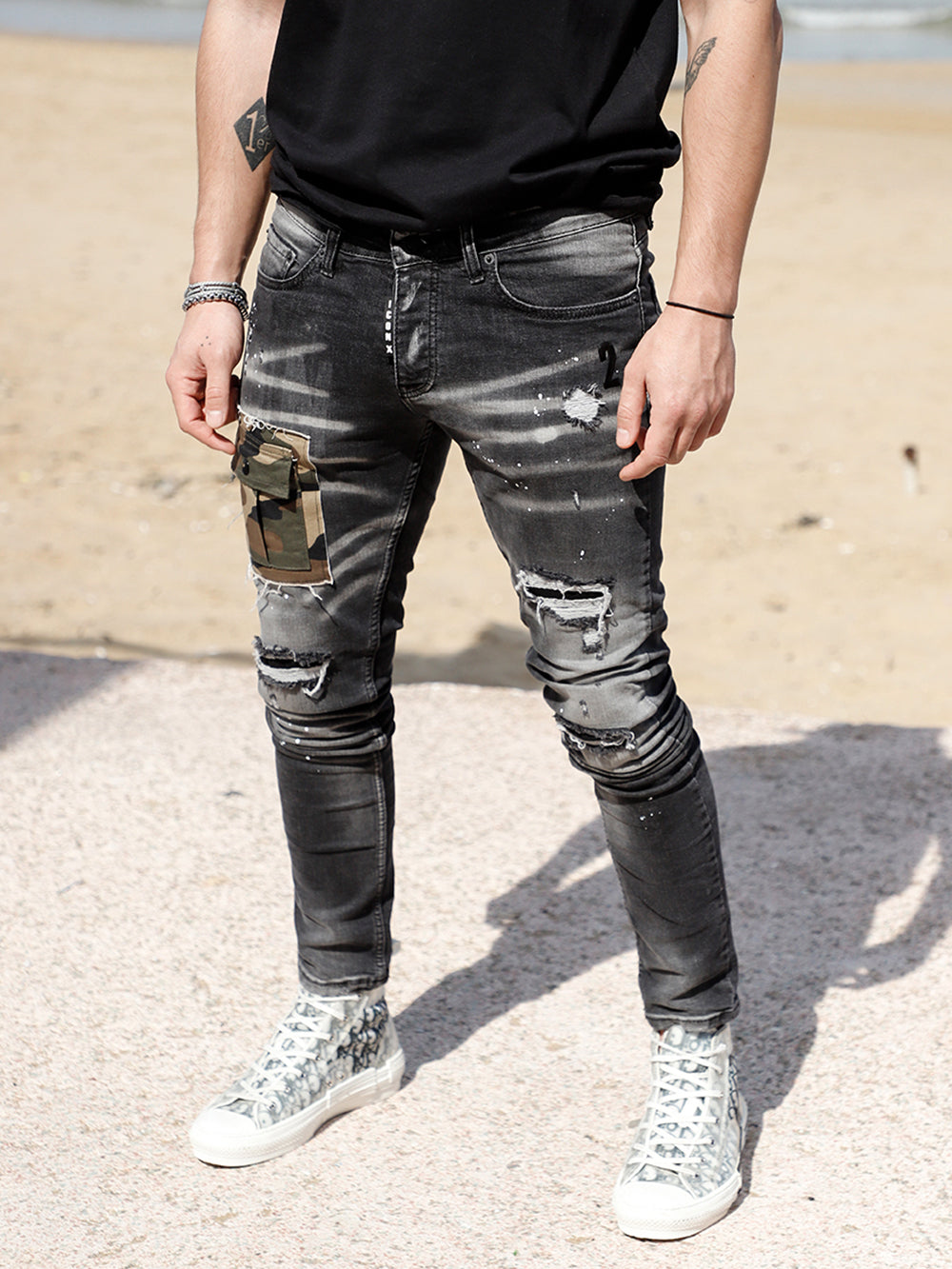 A man wearing a CAMOUFLAGE | BLACK t-shirt and jeans standing on the beach.