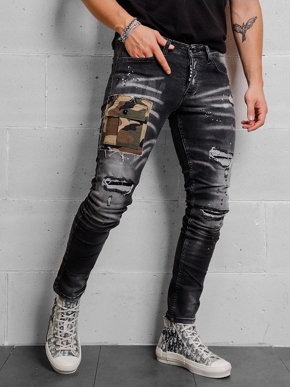 A man wearing ripped jeans and sneakers CAMOUFLAGE | BLACK leaning against a wall.