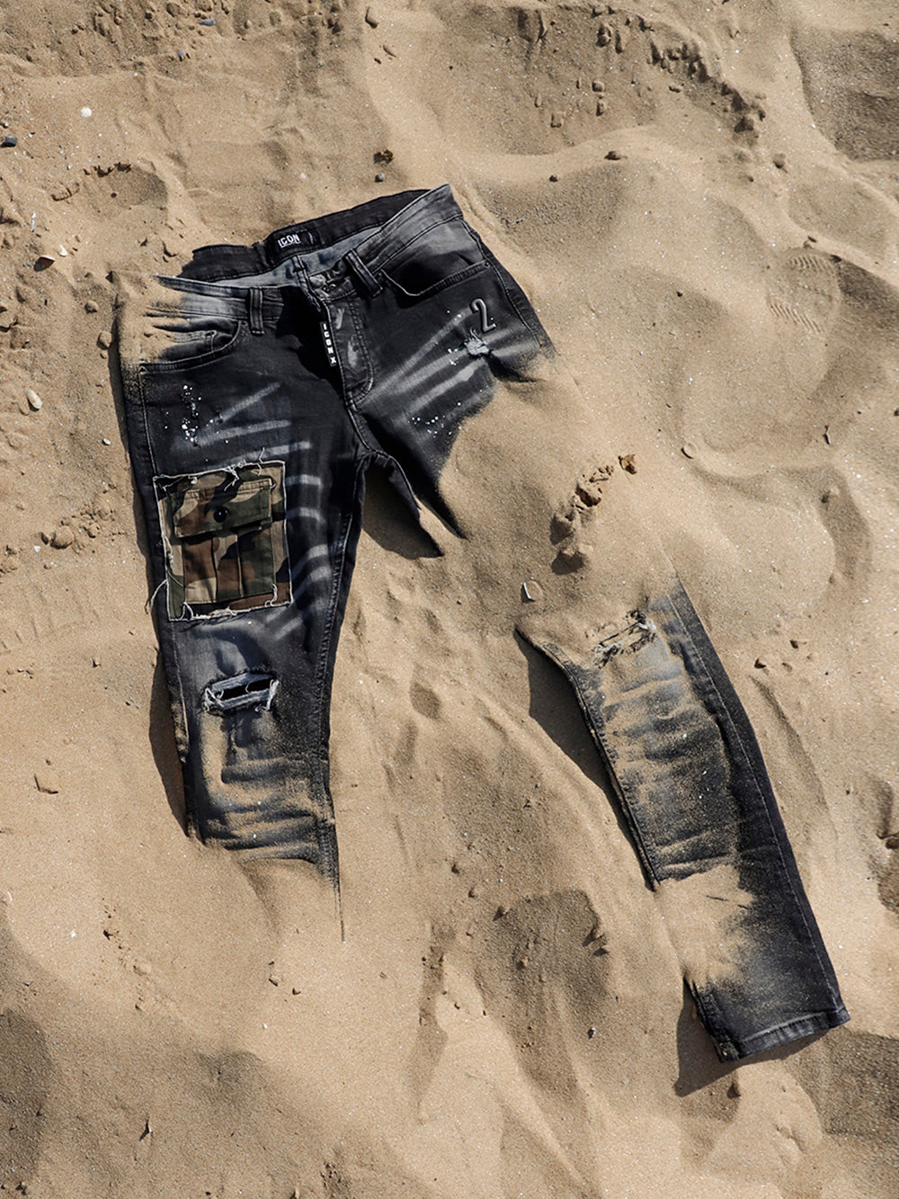 A pair of CAMOUFLAGE | BLACK jeans laying in the sand.