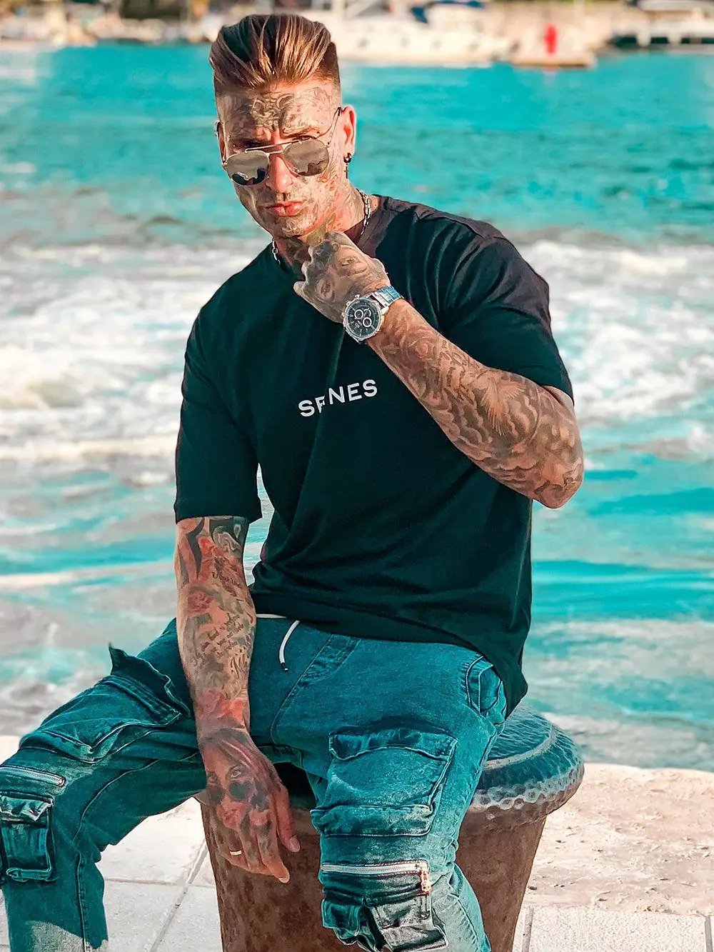 A man with tattoos and ripped jeans wearing THE ORIGINAL T-SHIRT sitting on a bench next to the ocean.