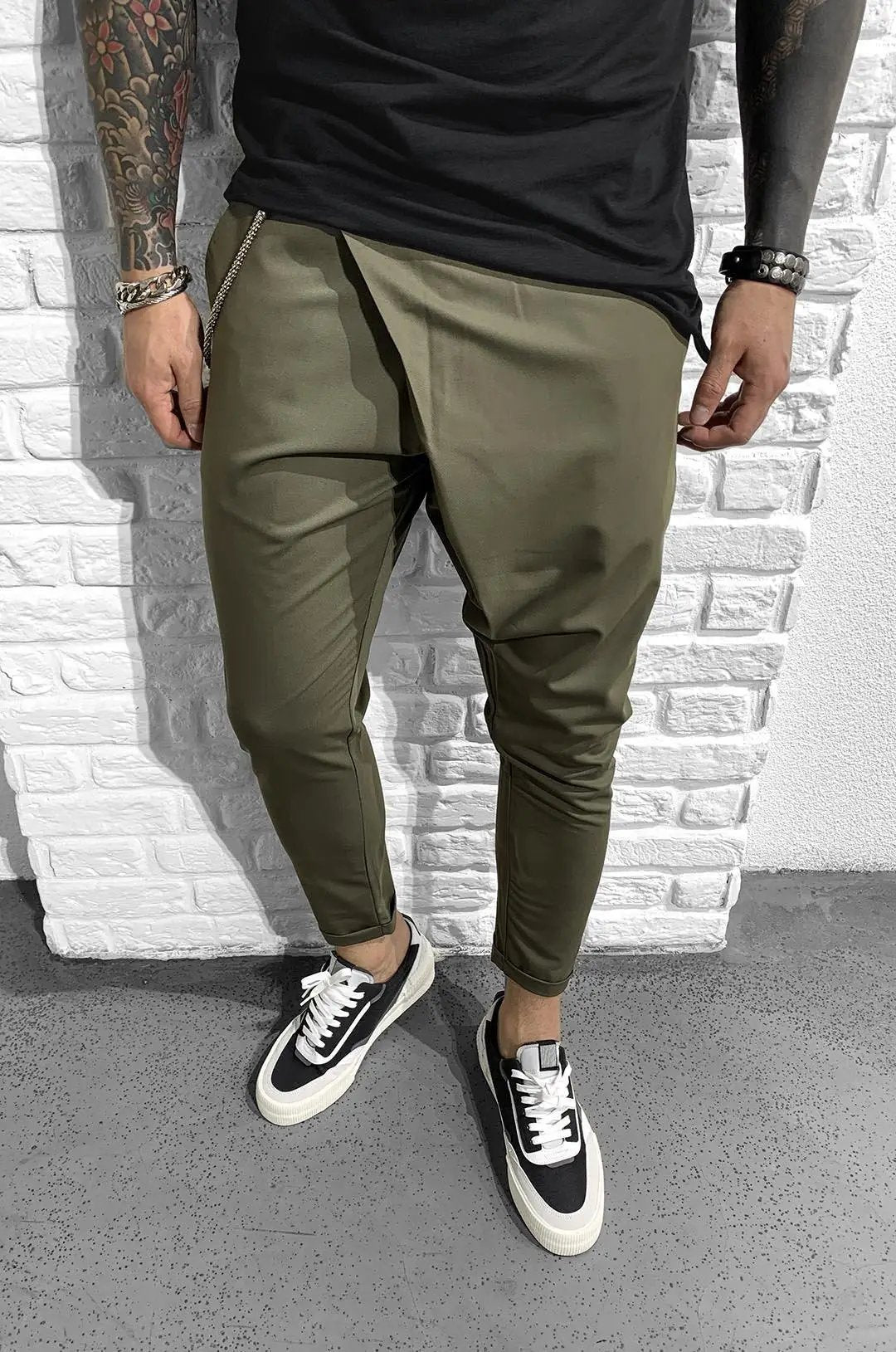 A man in ASSYMMETRIC ROLL UP - KHAKI pants with a comfortable fit standing next to a brick wall.