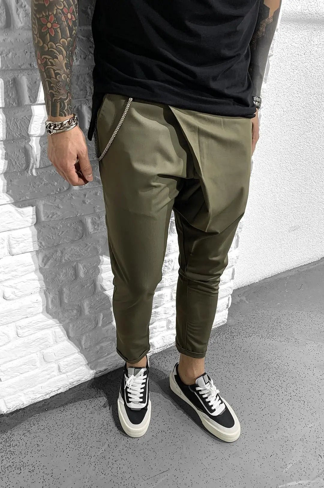 A man with tattoos is standing next to a wall, wearing clothes made from ASSYMMETRIC ROLL UP - KHAKI fabric for a comfortable fit and smooth feel.