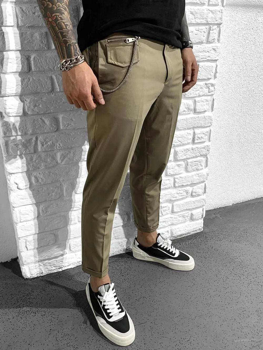 A man wearing Gentleman's Roll Up pants is standing next to a brick wall.