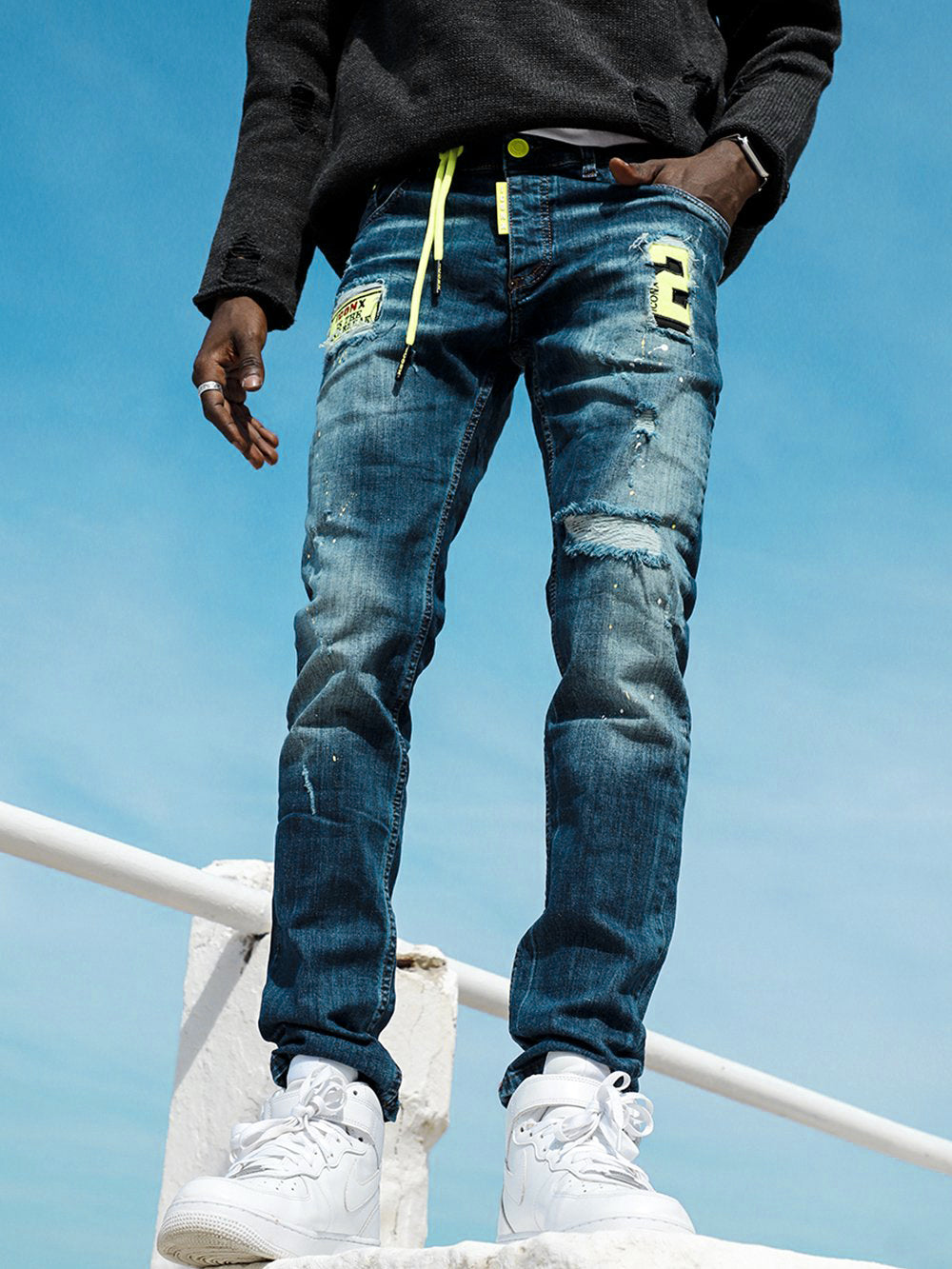 A man is standing on a ledge wearing PHOSPHORUS jeans and sneakers.